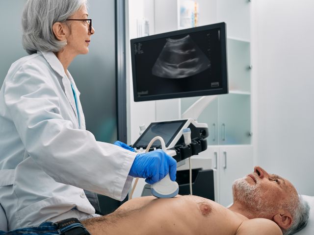 Ärztin führt eine Ultraschalluntersuchung am Brustkorb eines älteren Mannes durch, Ultraschallbild auf Monitor sichtbar.