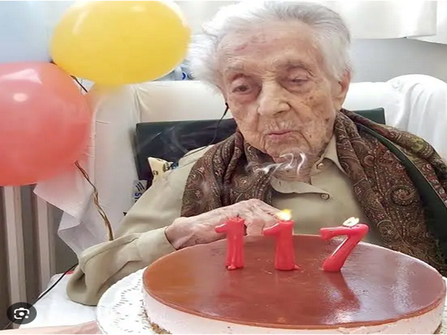 Ältere Person feiert Geburtstag mit einer Torte und Kerzen in Form der Zahl 117, bunte Luftballons im Hintergrund